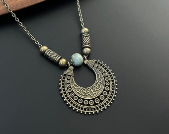 May include: A silver necklace with a large, ornate pendant shaped like a crescent moon. The pendant has a detailed design with circles and swirls. The necklace also features smaller silver beads.