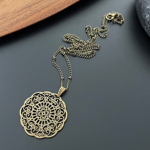 Peut inclure: Un collier doré avec un grand pendentif circulaire complexe. Le pendentif est en métal filigrané et présente un motif délicat et ajouré.