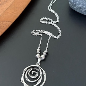 May include: A silver-toned pendant necklace featuring a spiral design. The pendant hangs from a delicate chain, with a cluster of small beads near the clasp. The necklace is displayed on a black surface.