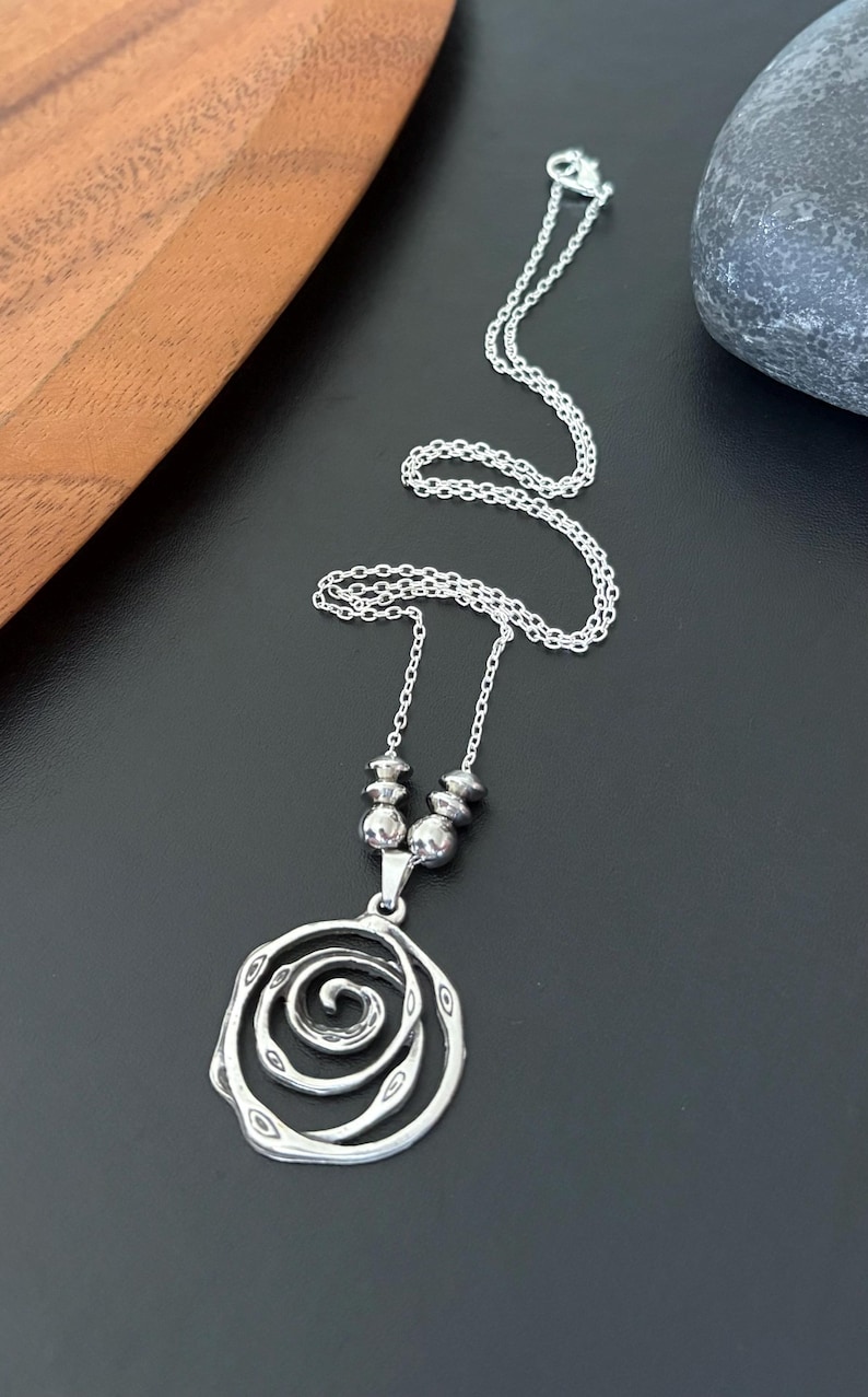 May include: A silver-toned pendant necklace featuring a spiral design. The pendant hangs from a delicate chain, with a cluster of small beads near the clasp. The necklace is displayed on a black surface.