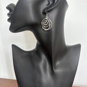 May include: Silver-toned spiral earrings. The earrings are displayed on a black mannequin head. The design features concentric circles, creating a textured, openwork appearance. The earrings hang from a simple hook.