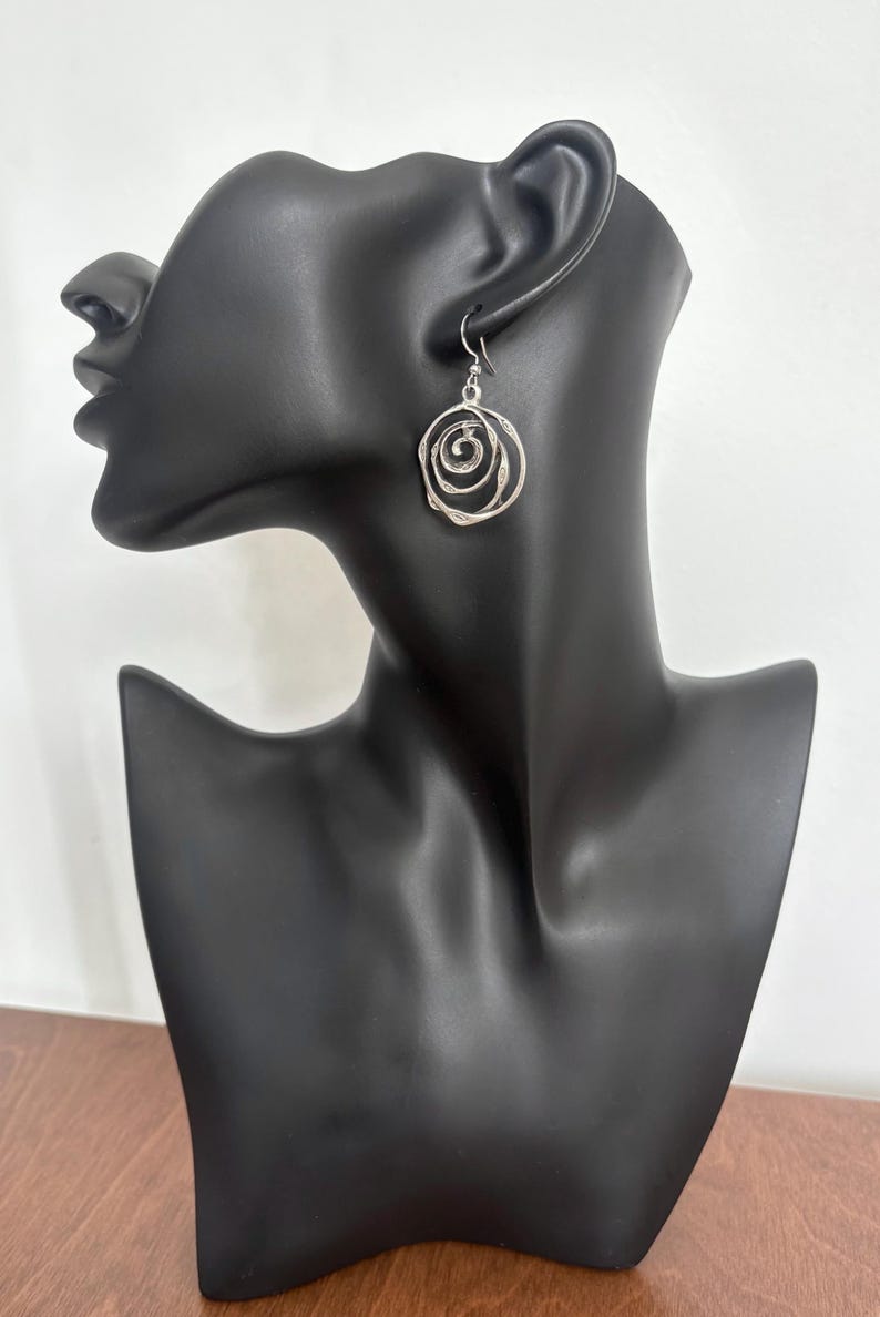 May include: Silver-toned spiral earrings. The earrings are displayed on a black mannequin head. The design features concentric circles, creating a textured, openwork appearance. The earrings hang from a simple hook.