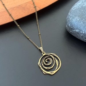 Peut inclure: Un collier pendentif doré avec un motif en spirale. Le pendentif est suspendu à une chaîne délicate avec de petites perles rondes. Le collier est présenté sur une surface sombre, avec un élément en bois et une pierre grise en arrière-plan.