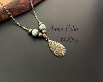 May include: A bronze necklace with a teardrop-shaped pendant and white beads. The pendant has a textured design. The necklace is on a dark background. Aspen Boho @Etsy