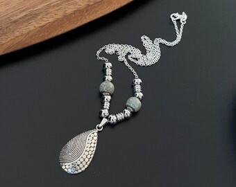 May include: Silver-toned necklace with a teardrop pendant. The pendant features a textured design with a combination of lines and small circles. The necklace includes two gray stone beads and a delicate chain. The necklace is displayed on a black surface.