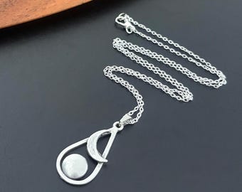 May include: A silver-colored necklace with a pendant featuring a crescent moon and a circular design. The pendant hangs from a delicate chain. The necklace is displayed on a dark surface.