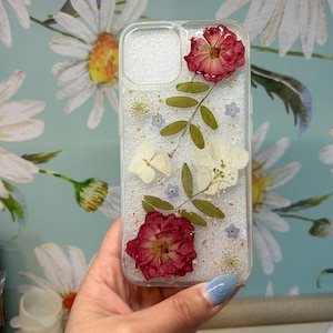 May include: A clear phone case with dried flowers and gold leaf accents. The flowers include red roses, white flowers, and blue forget-me-nots.