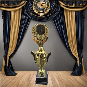 Puede incluir: Un trofeo dorado con una base negra y un diseño de corona de laurel. El trofeo tiene una parte superior circular con un centro negro.