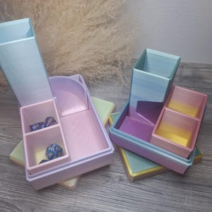 Könnte beinhalten: Zwei pastellfarbene Würfelbehälter, einer offen und einer geschlossen, mit mehreren Fächern. Das offene Tablett hat Würfel im Inneren. Die Tabletts sind in Blau-, Rosa-, Lila- und Gelbtönen gehalten. Die Tabletts stehen auf einer hellgrünen und gelben Oberfläche.