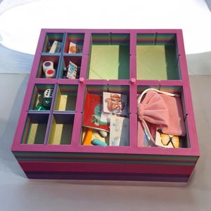 Könnte beinhalten: Ein quadratischer Aufbewahrungs-Organizer mit mehreren Fächern, der durch ein regenbogenfarbenes Streifenmuster an der Außenseite und den Innenwänden besticht. Die Box enthält diverse Kleinteile wie Würfel, einen rosa Samtbeutel und Karten.