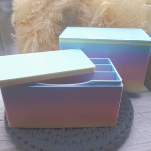 Könnte beinhalten: Zwei rechteckige Aufbewahrungsboxen mit Deckeln. Die Boxen haben eine Farbverlaufsoptik, die von Rosa über Lila zu Blau übergeht. Die kleinere Box hat einen offenen Deckel, der Innenfächer freilegt. Die Boxen stehen auf einer grauen Häkelmatte.
