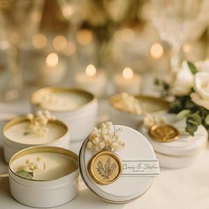 May include: White candles in round tins with gold rims and lids. Each candle is decorated with dried flowers and a gold seal. One lid has the text "Anne & Stefan".