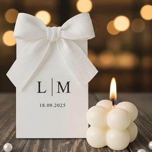 Puede incluir: Una caja de regalo blanca con un gran lazo blanco, las iniciales "L | M" y la fecha "18.09.2025" impresas en la parte delantera. Una vela de burbujas blanca encendida está colocada junto a la caja. El fondo está borroso con luces bokeh.