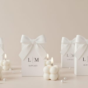 May include: A collection of white bubble candles with lit flames, alongside white gift boxes adorned with bows. The boxes display initials and a date. Scattered pearls provide a decorative element to the arrangement.