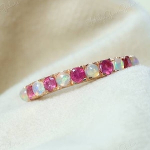 May include: A delicate ring featuring alternating round-cut gemstones. The ring showcases vibrant pink rubies and iridescent opals set in a gold band. The gemstones are evenly spaced, creating a continuous pattern. The ring is displayed on a soft, white fabric.