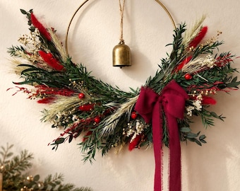 May include: A circular gold metal hoop wreath adorned with dark green foliage, red dried grasses, white filler flowers, and small red berries. A brass bell hangs from the top center, and a deep red fabric bow with long trailing ribbons is attached.