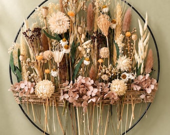 May include: A circular wall hanging featuring a black metal hoop filled with an arrangement of dried flowers and grasses in shades of cream, beige, tan, and dusty pink. A horizontal jute twine band secures the stems across the lower section.