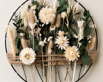 May include: A circular black metal hoop wall hanging featuring an arrangement of dried flowers and greenery. The stems are secured horizontally across the lower half of the hoop with natural jute twine, showcasing white daisies, grasses, and eucalyptus.