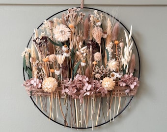May include: A circular floral wall hanging featuring dried flowers in shades of cream, beige, and pink. The arrangement includes various textures and shapes, set within a black metal hoop. The base of the arrangement has a woven design.
