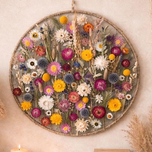 May include: A circular wall hanging featuring a wire mesh frame filled with an assortment of dried flowers in shades of yellow, pink, purple, and white. The arrangement includes various blossoms, grasses, and seed pods secured within the frame.