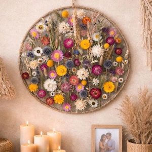 May include: A circular floral wall hanging with a variety of dried flowers in shades of yellow, pink, purple, and white. The arrangement is set against a neutral wall, with candles and a wooden table in the foreground. A woven lamp and macrame wall decor are also visible.