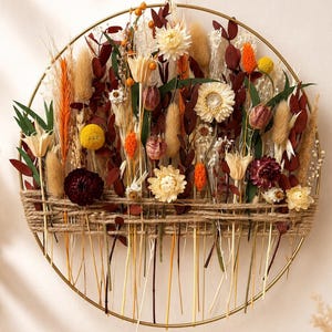 Pode incluir: Uma decoração de parede circular em metal dourado com um arranjo de flores e ervas secas em tons de creme, laranja, amarelo e vermelho escuro. Os caules estão presos à metade inferior do aro com um cordão de juta grosso de cor natural.