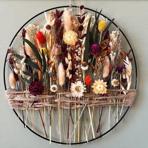 Pode incluir: Decoração de parede circular com flores secas em tons de vermelho, amarelo, branco e roxo. As flores estão dispostas dentro de um aro de metal preto, com uma base de barbante trançado. O design é natural e rústico.