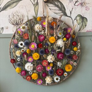 May include: A circular floral wall hanging featuring a variety of dried flowers in shades of yellow, pink, red, and white. The flowers are arranged within a woven, rope-like frame. A natural, decorative piece for home decor.