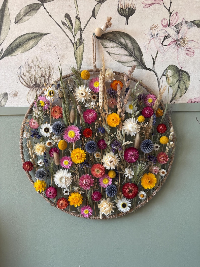 May include: A circular floral wall hanging featuring a variety of dried flowers in shades of yellow, pink, red, and white. The flowers are arranged within a woven, rope-like frame. A natural, decorative piece for home decor.