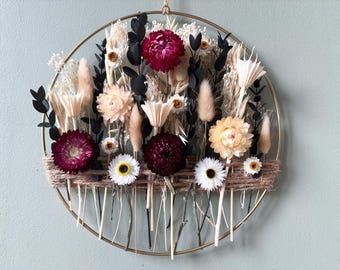 May include: A circular floral wall hanging with a gold metal ring. The arrangement features dried flowers in shades of burgundy, cream, and white, with black accents. A woven twine element adds texture. The overall design is natural and decorative.