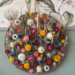 May include: A circular floral wall hanging featuring a variety of dried flowers in shades of yellow, pink, red, and white. The flowers are arranged within a woven frame, creating a natural and decorative piece. A jute rope is used for hanging.