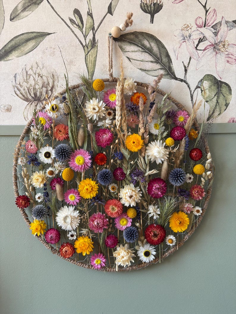 May include: A circular floral wall hanging featuring a variety of dried flowers in shades of yellow, pink, red, and white. The flowers are arranged within a woven frame, creating a natural and decorative piece. A jute rope is used for hanging.
