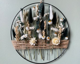 May include: A circular wall hanging featuring dried flowers and greenery. The arrangement includes white daisies, fluffy white plumes, and dark green accents, all set against a black metal ring. A woven jute band adds texture.
