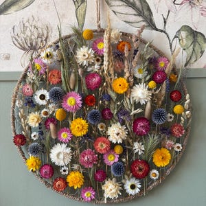May include: A circular floral wall hanging featuring a variety of dried flowers in shades of yellow, pink, red, and white. The flowers are arranged within a woven, circular frame, creating a natural and decorative piece. The background features a floral print.