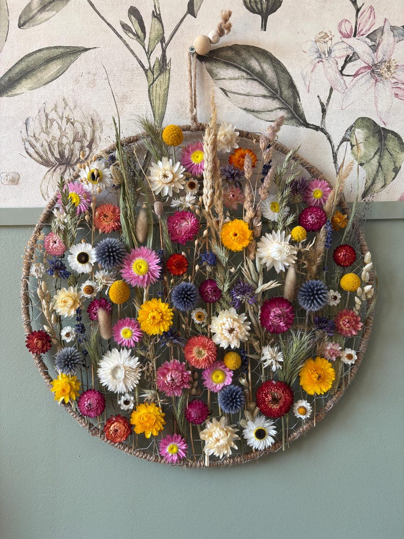 May include: A circular floral wall hanging featuring a variety of dried flowers in shades of yellow, pink, red, and white. The flowers are arranged within a woven, circular frame, creating a natural and decorative piece. The background features a floral print.