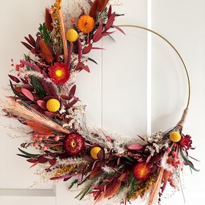 Può includere: Una ghirlanda a semicerchio con fiori secchi nei toni del rosso, arancione, giallo e verde. I fiori sono disposti su un cerchio di metallo dorato, con una sezione avvolta in spago. Un elemento decorativo.