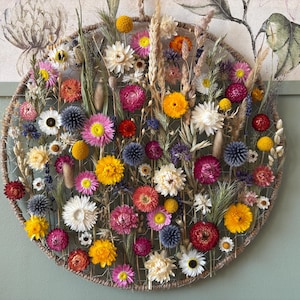 May include: A circular floral arrangement featuring a variety of dried flowers in shades of yellow, pink, white, and blue. The arrangement is set against a neutral background, creating a visually appealing display. The flowers are arranged within a woven circular frame.