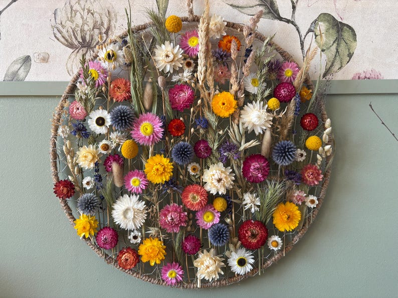 May include: A circular floral arrangement featuring a variety of dried flowers in shades of yellow, pink, white, and blue. The arrangement is set against a neutral background, creating a visually appealing display. The flowers are arranged within a woven circular frame.