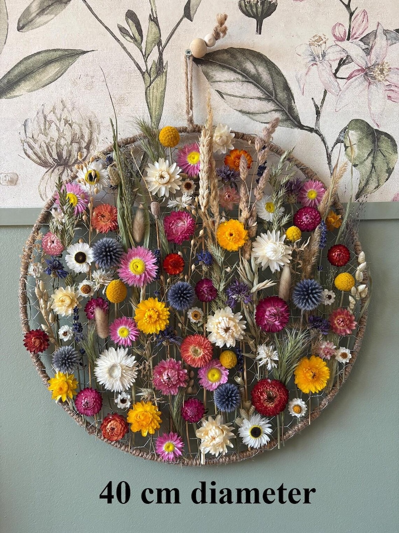 May include: A circular floral wall hanging, 40 cm in diameter, featuring a variety of dried flowers in shades of yellow, pink, orange, and white. The flowers are arranged within a woven frame, creating a natural and decorative piece. The text "40 cm diameter" is at the bottom.