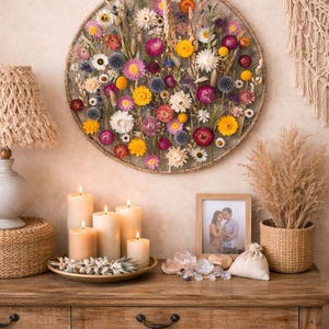 May include: A circular floral wall hanging with a variety of dried flowers in shades of yellow, pink, purple, and white. The arrangement is set against a neutral wall, with candles and a wooden table in the foreground. A woven lamp and macrame wall decor are also visible.
