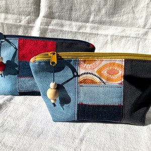 Könnte beinhalten: Zwei Jeans-Patchwork-Reißverschlusstaschen mit Holzperlen-Akzenten. Eine Tasche hat einen roten Akzent, die andere ein orange-weißes Muster. Beide haben goldfarbene Reißverschlüsse und liegen auf einer weißen Oberfläche.