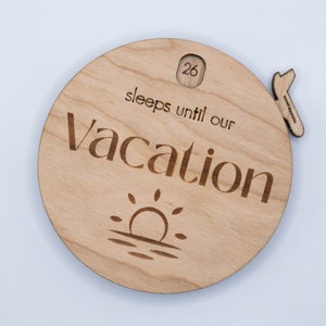 May include: A circular wooden countdown sign, engraved with the words "sleeps until our Vacation". It features a sun and water graphic, and a small wooden piece with the number 26. The sign is made of light-colored wood.