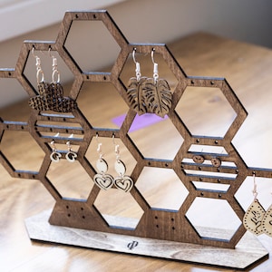 May include: Wooden hexagonal earring display stand featuring a selection of earrings, including leaf-shaped, heart-shaped, and geometric designs. The stand has a brown finish and a small inscription on the base.