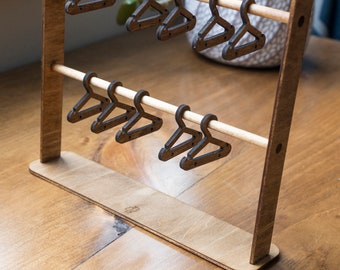 May include: A wooden dollhouse-sized clothing rack with two wooden dowels and eight miniature wooden hangers. The rack is made of light brown wood and the hangers are dark brown.