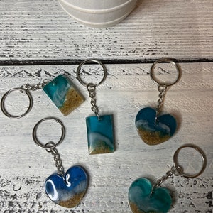 May include: Five resin keychains with ocean-themed designs. Each keychain features a different color combination of blue, green, and white with a layer of sand at the bottom. The keychains are attached to silver key rings.