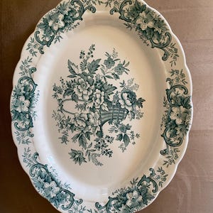 May include: Oval serving platter with a white base and a teal floral design. The platter features a scalloped edge and a central floral basket motif, surrounded by floral and scrollwork accents. A classic piece for serving.