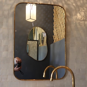 Pode incluir: Um espelho retangular com cantos arredondados e moldura dourada está pendurado acima de uma bacia de lavatório cor de pêssego. O espelho reflete uma luminária e um espelho menor, com formato único. Uma torneira dourada e uma pequena tigela dourada também são visíveis.