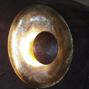 May include: A large, round, gold-coloured metal wall sconce with a textured surface. The sconce features a dark, circular centre and emits a warm, yellow light. The design is modern and decorative, suitable for interior lighting.