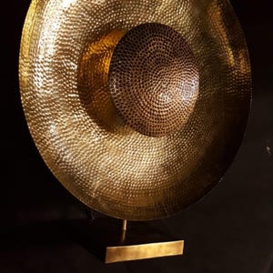 May include: A decorative, hammered brass sculpture. It features a large, circular outer element and a smaller, central, spherical component. The sculpture is mounted on a rectangular base with a single, thin support, all in a warm, golden hue. The surface has a dimpled pattern.