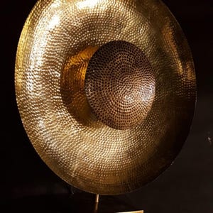May include: A large, hammered brass gong with a central, smaller, circular element. The gong has a warm, golden hue and is mounted on a slender, gold-coloured stand. The surface is textured with a pattern of small, evenly spaced indentations.